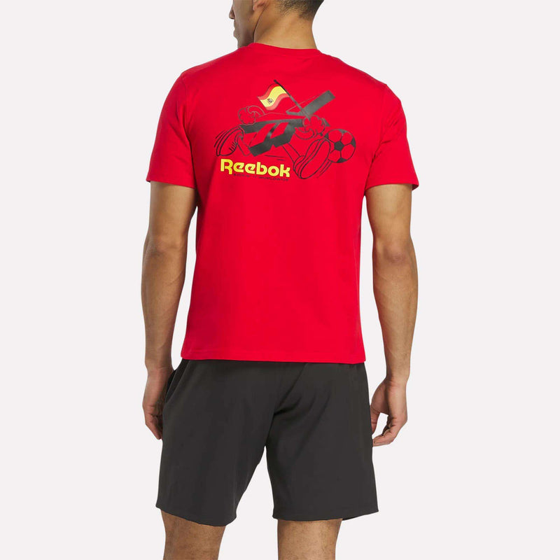 Someone stands facing away from the camera wearing a bright red Reebok España Football T-Shirt featuring a cartoon graphic and logo on the back, paired with black shorts.