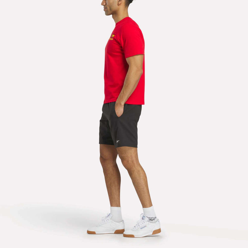 A person stands in profile against a plain white background, wearing an España Football T-Shirt and white Reebok sneakers, paired with black shorts and white socks.