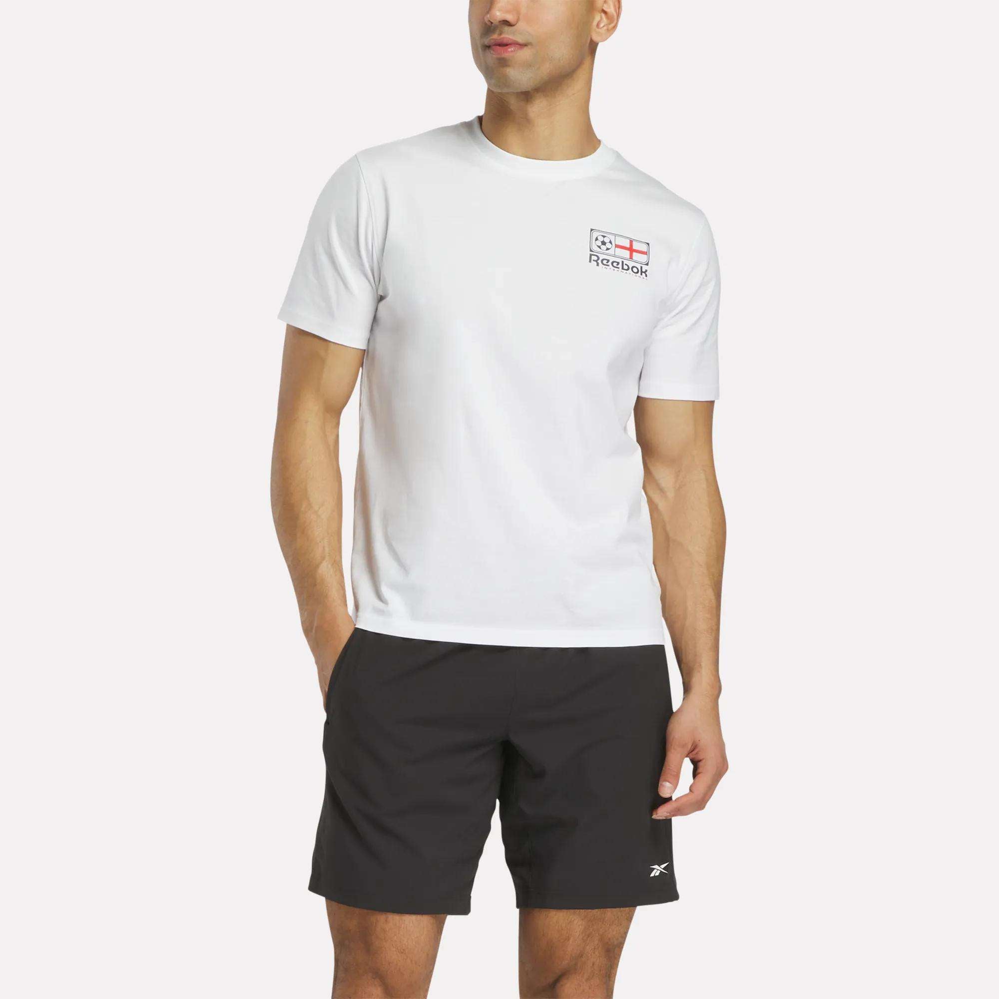 A man wears a white Reebok England Football T-Shirt with a small logo and flag on the chest, and black Reebok shorts, standing with one hand in his pocket against a plain background.