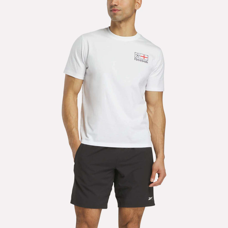 A man wears a white Reebok England Football T-Shirt with a small logo and flag on the chest, and black Reebok shorts, standing with one hand in his pocket against a plain background.