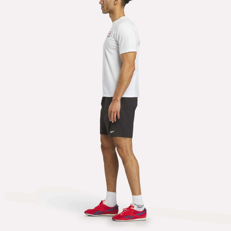 A man stands in profile against a plain white background, wearing a white Reebok England Football T-Shirt, black athletic shorts, white socks, and red sneakers.