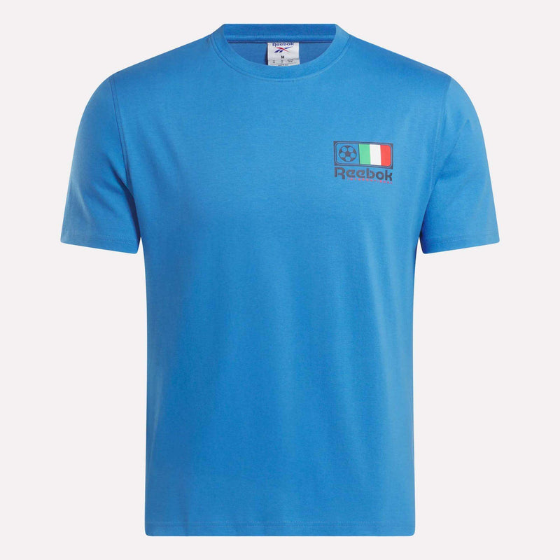 The Reebok Italia Football T-Shirt is blue, made from soft cotton jersey with short sleeves. A small soccer ball, Italian flag, and black Reebok logo decorate the left chest. Displayed on a plain white background.