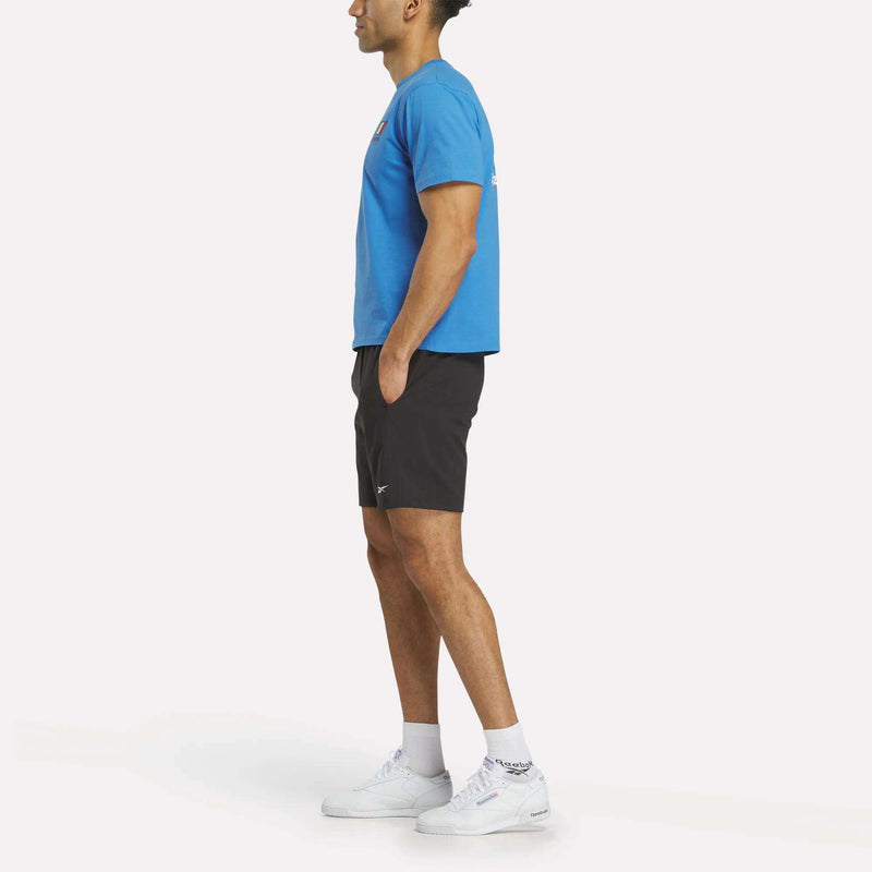 A person stands in profile, hands in pockets, wearing a blue Reebok Italia Football T-Shirt, black shorts, white socks, and white sneakers against a plain white background.
