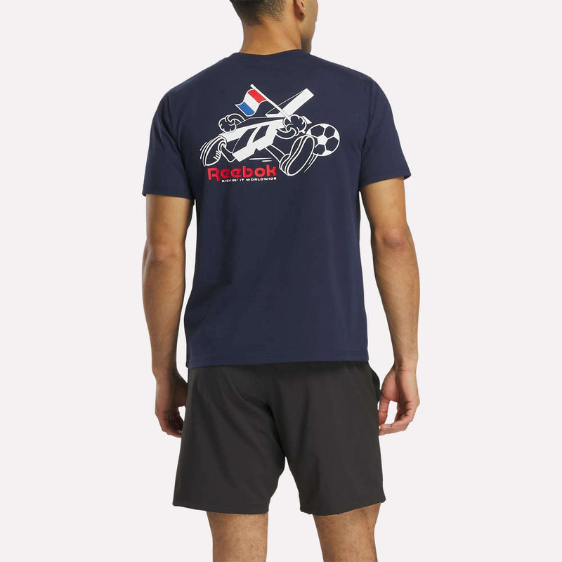 A man faces away, wearing a Reebok France Football T-Shirt with graphics and black shorts. The shirt's back features illustrations of a baseball bat, ball, tennis racket, and football.