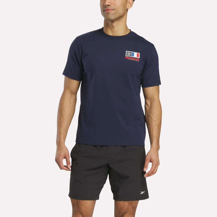 A man wears a Reebok France Football T-Shirt in navy blue, featuring a small graphic and text on the left chest, paired with black shorts, standing against a plain white background.