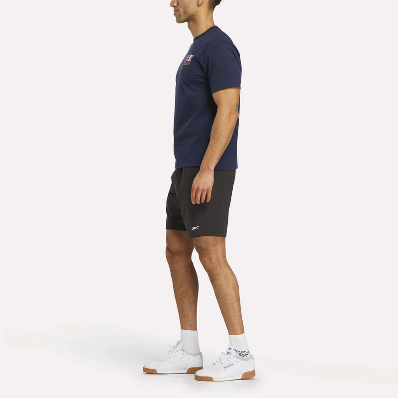 A man stands sideways against a plain white background, wearing a navy Reebok France Football T-Shirt, black athletic shorts, white crew socks, and white sneakers.