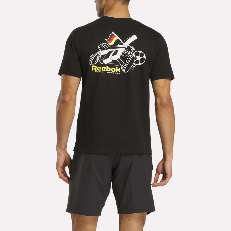 A man stands facing away, wearing a black Reebok Deutschland Football T-Shirt featuring sports equipment graphics and Reebok text on the back, paired with dark shorts.