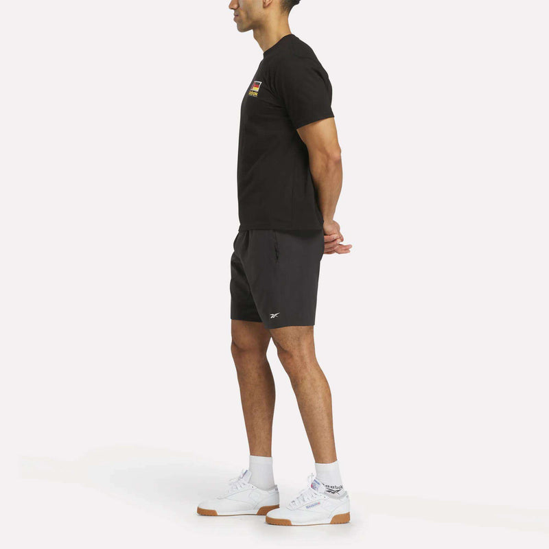 A person stands in profile against a plain background, wearing the Reebok Deutschland Football T-Shirt, black shorts, and white Reebok sneakers. Their hands are clasped behind their back as they look forward.