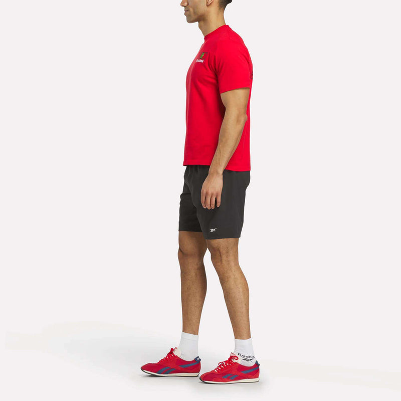 A man stands in profile wearing a red Reebok Portugal Football T-Shirt, black athletic shorts, white socks, and red sneakers against a plain white background.