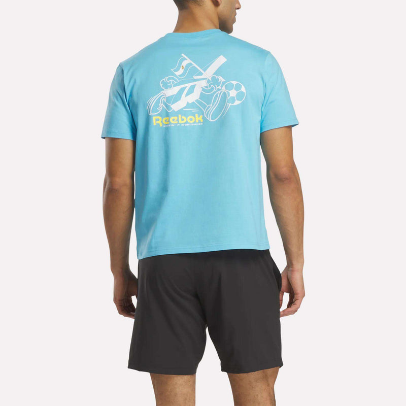 A man faces away wearing a light blue Reebok Argentina Football T-Shirt with white sports equipment graphics and a yellow Reebok logo on the back, paired with black shorts.