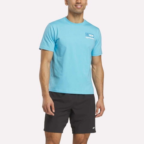 A man wearing a light blue Reebok Argentina Football T-Shirt and black shorts stands against a plain white background, smiling with his arms relaxed at his sides.