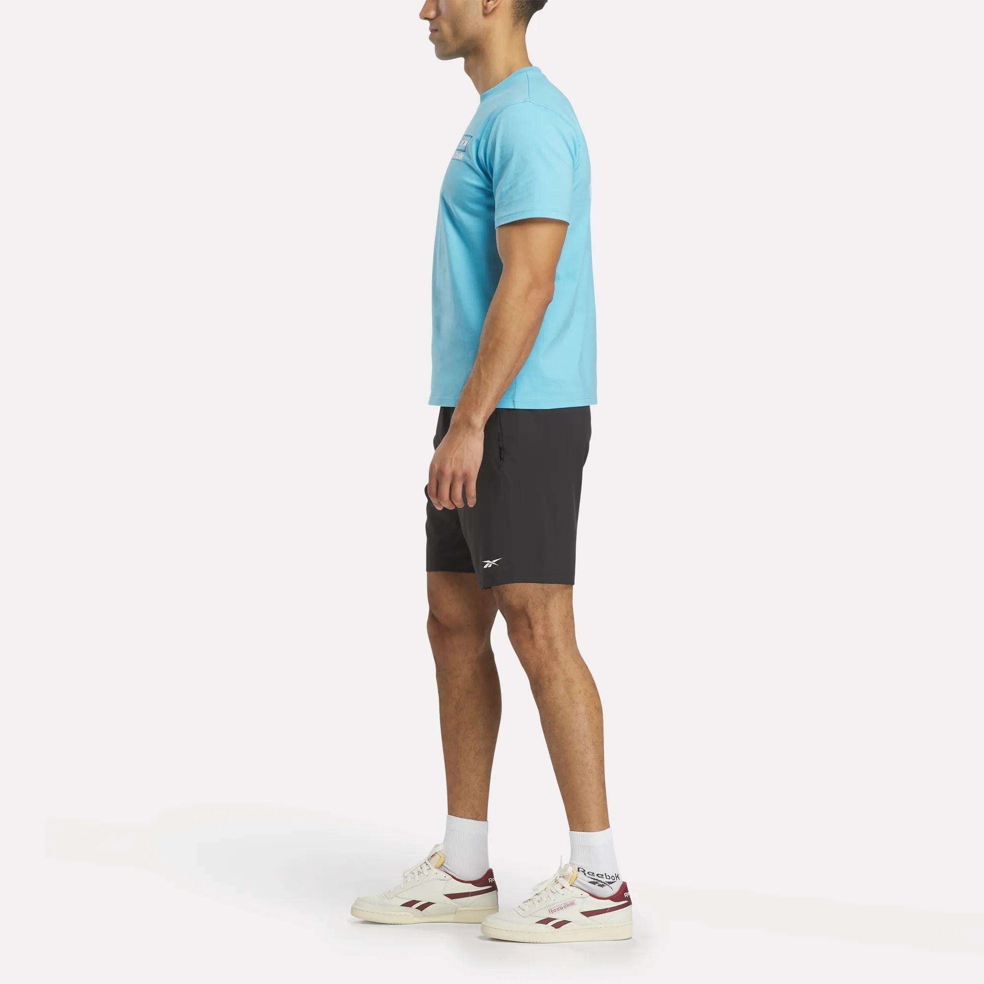 A man in profile wears a light blue Argentina Football T-Shirt and black shorts, paired with white Reebok sneakers with red accents, all set against a plain white background.