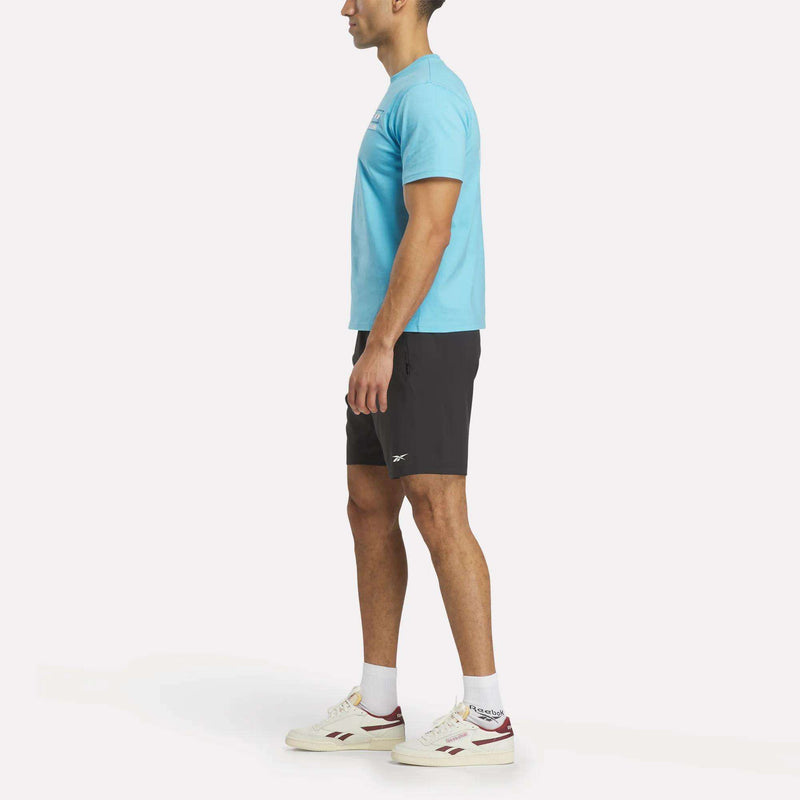 A man in profile wears a light blue Argentina Football T-Shirt and black shorts, paired with white Reebok sneakers with red accents, all set against a plain white background.