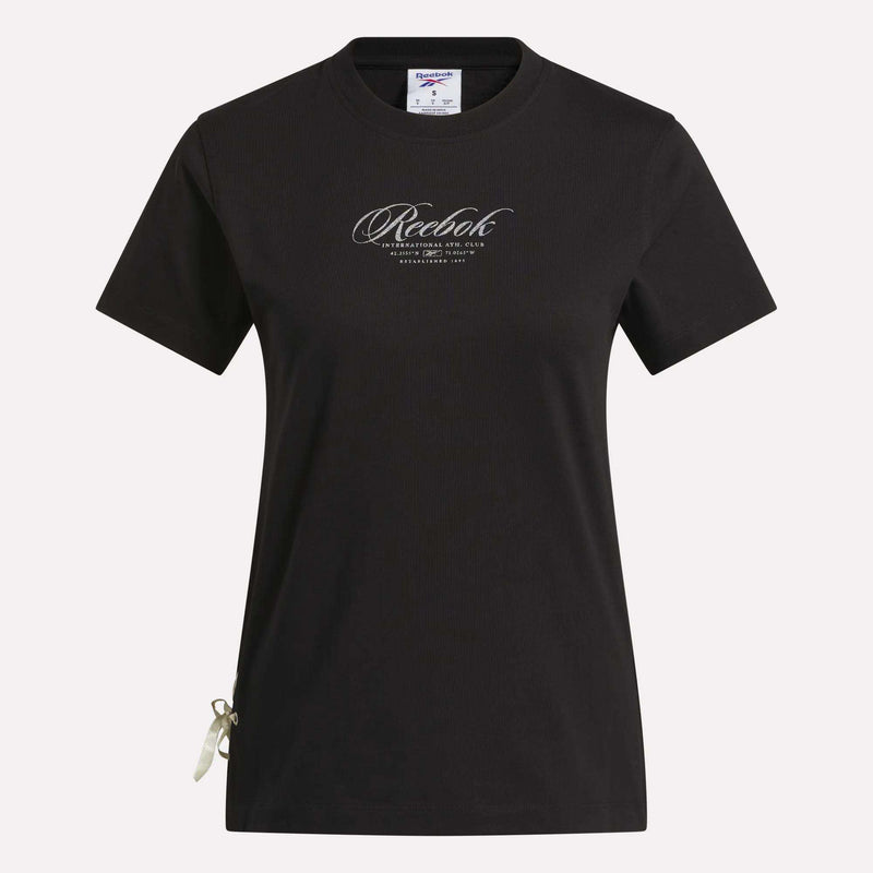 The Reebok Women's Ribbon T-Shirt is shown in black with short sleeves against a white background. It features the brand’s cursive logo on the chest and a small ribbon detail at the bottom left hem. Made from soft cotton, this t-shirt offers comfortable wear and metallic shine for a stylish everyday look.
