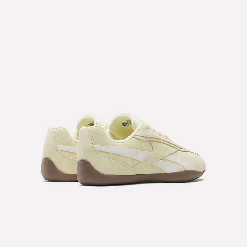 A pair of Reebok Women’s Ultra Lo Shoes in light yellow, featuring white stripes, a suede upper, and brown soles are displayed at an angle against a plain white background.