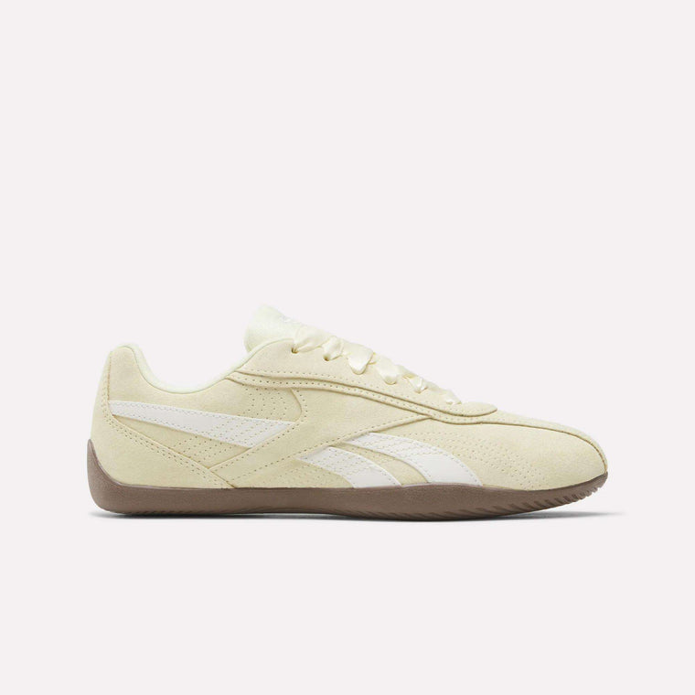 The Reebok Women’s Ultra Lo Shoes, shown here in Raw Yellow/Chalk/Gum, feature a beige suede upper accented with white stripes and laces, perforated details, and a brown rubber sole—blending classic style with motorsport-inspired athletic flair in a side profile view against a plain white background.
