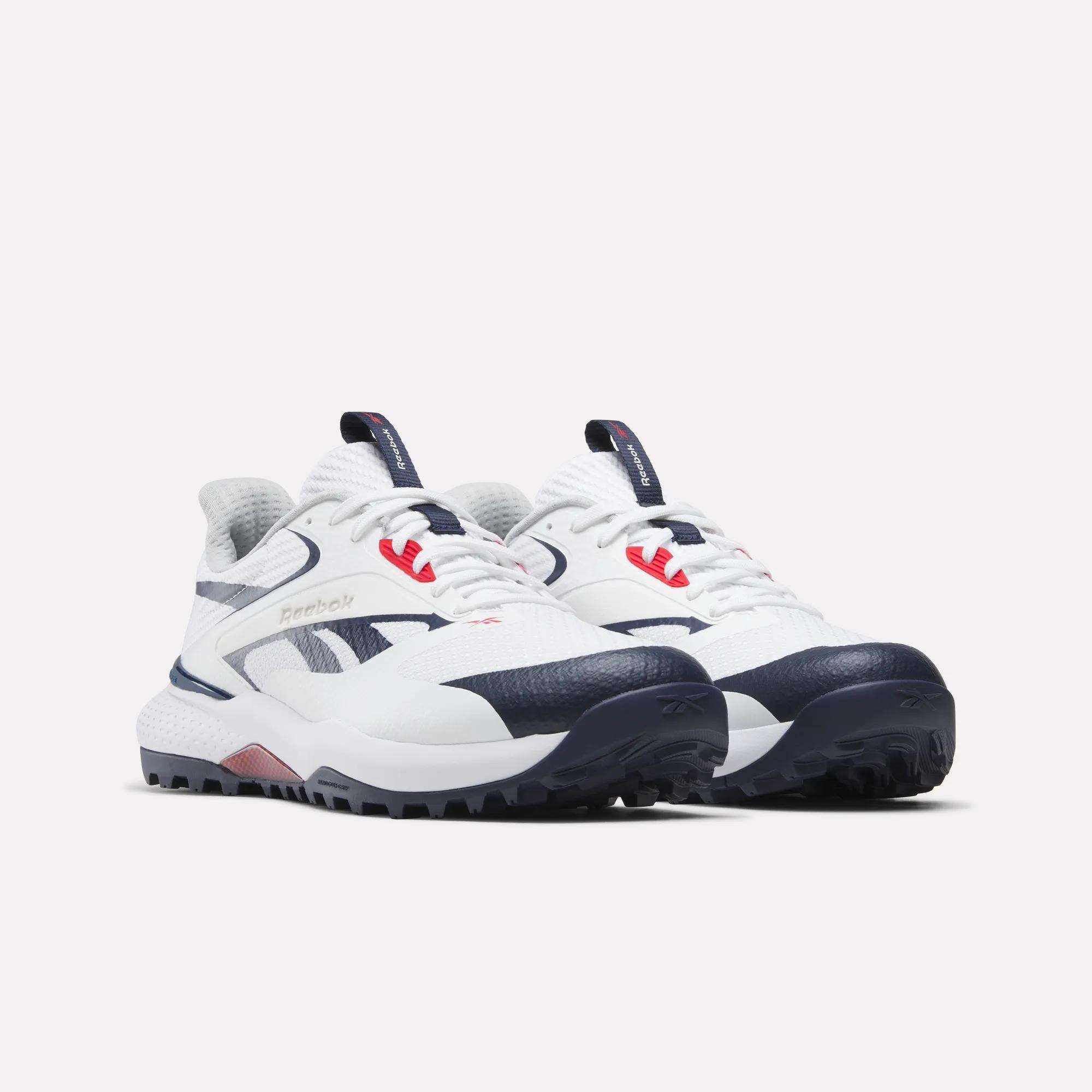 A pair of Reebok Nano Golf Shoes in white with navy blue and red accents, FlexWeave uppers, and rugged soles with Floatride foam, placed side by side on a light background.