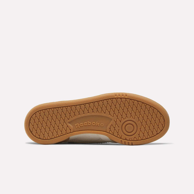 The image displays the sole of the Reebok Women’s Club C Grounds 70 Shoes, featuring a brown rubber outsole with a herringbone tread pattern and the Reebok logo embossed in the center. This detail enhances the classic charm of these retro sneakers.