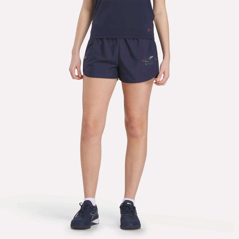 A person wearing a navy athletic outfit and Women's Reebok x F45 Training Coach Lux Shorts stands against a plain light background. Only the lower torso and legs are visible.