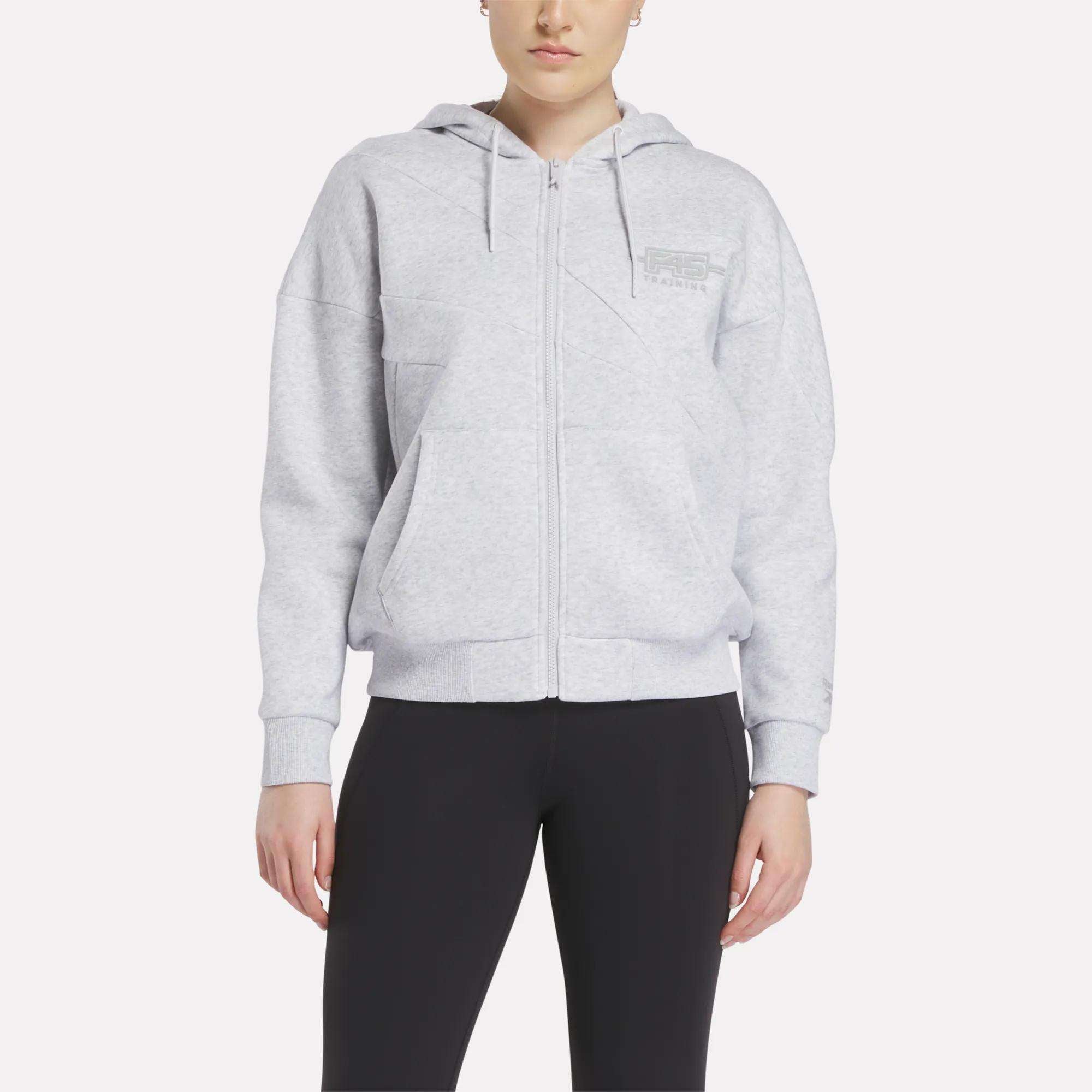 A woman models the Reebok x F45 Training Relaxed Zip Hoodie in light gray with a hood and subtle chest text, paired with black leggings, against a white background.