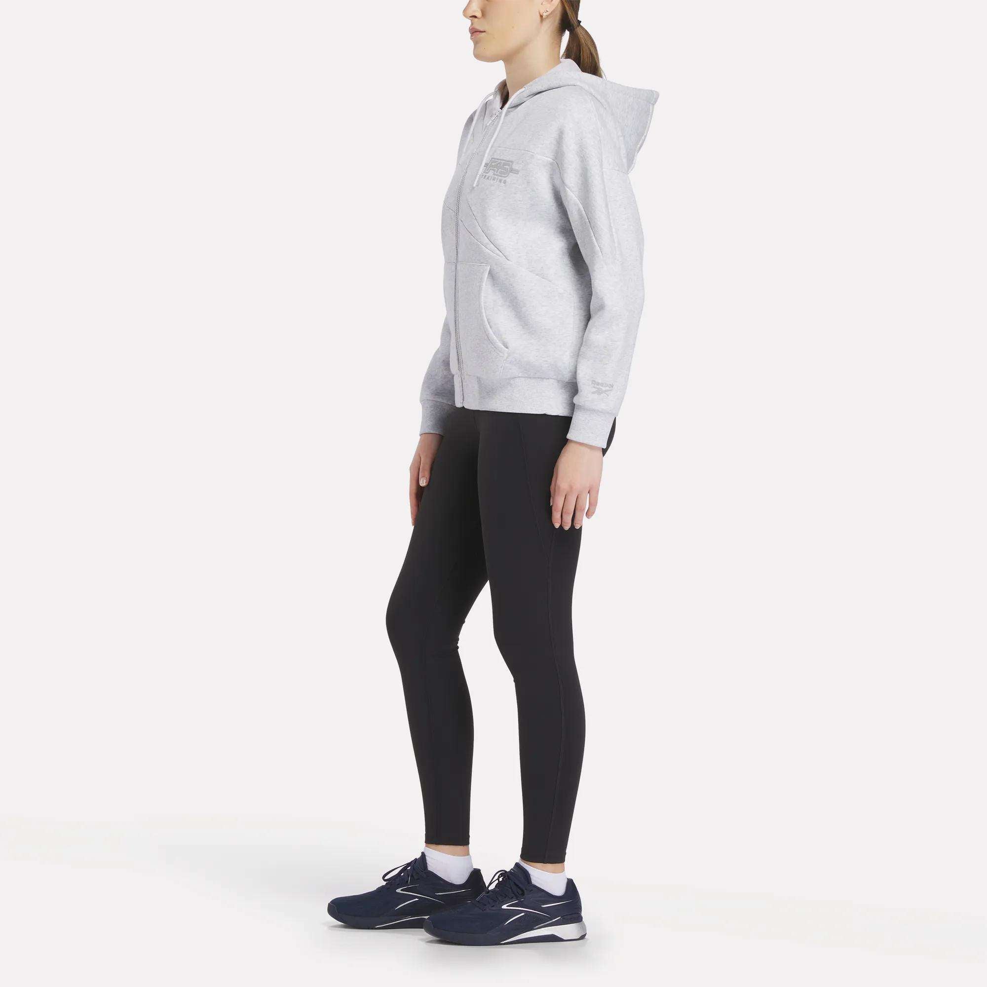A woman stands sideways in a light gray Women's Reebok x F45 Training Relaxed Zip Hoodie by Reebok, black leggings, and navy athletic shoes against a plain white background.