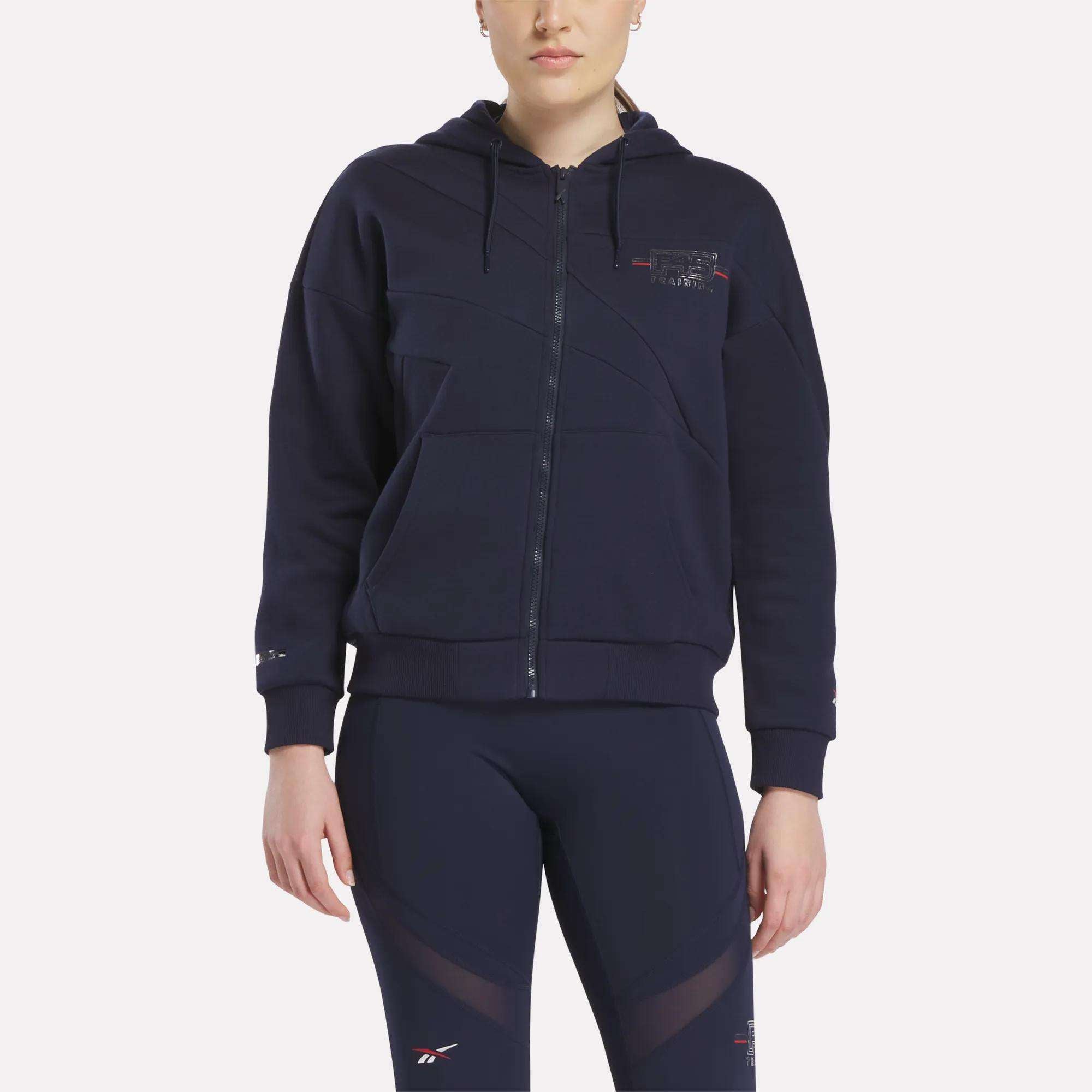 A woman stands facing forward in a Women's Reebok x F45 Coach Full-Zip Hoodie with a hood and navy leggings, both featuring subtle Reebok logos, against a white backdrop—perfect for F45 Training or everyday comfort.