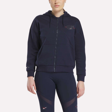 A woman stands facing forward in a Women's Reebok x F45 Coach Full-Zip Hoodie with a hood and navy leggings, both featuring subtle Reebok logos, against a white backdrop—perfect for F45 Training or everyday comfort.