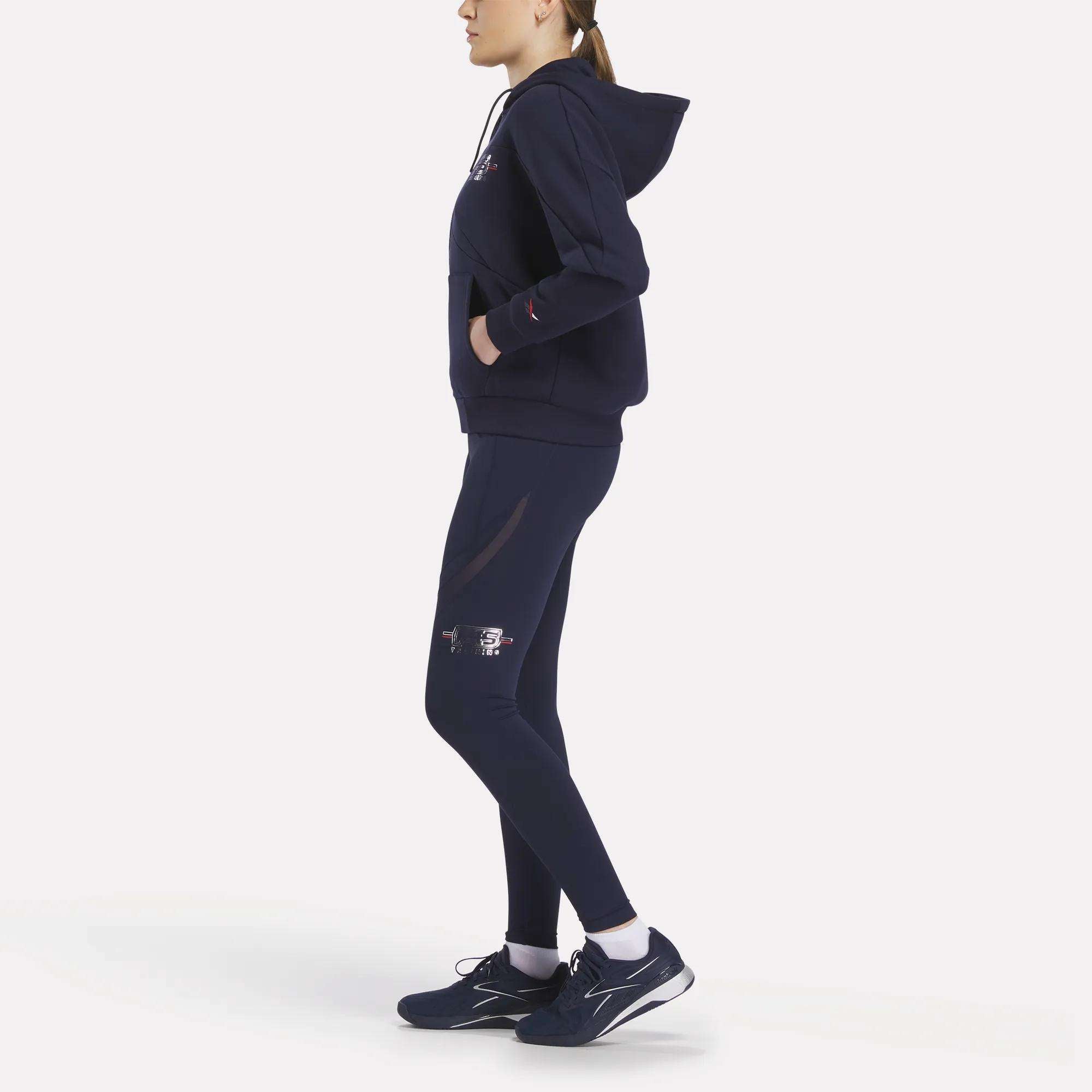 A woman stands in profile with hands in pockets, wearing a Women's Reebok x F45 Coach Full-Zip Hoodie, matching leggings, white socks, and navy athletic shoes. Her hair is tied back against a plain white background—perfect for F45 Training.