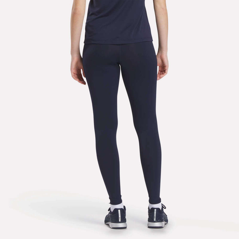 Person facing away from the camera wears a navy blue Reebok tee, Women's Reebok x F45 Training Coach Lux Leggings, white socks, and dark athletic shoes, all set against a plain light background.