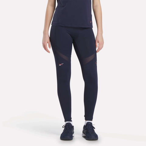 A woman in Reebok x F45 Training Coach Lux Leggings and a navy tee, both with subtle white logos and mesh panels, pairs the look with black athletic shoes—perfect for intense workouts—standing against a plain light background.