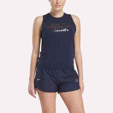 A woman wears the Women's Reebok x F45 Training Coach Tank Top in navy blue with matching shorts, both featuring subtle designs and small logo details, perfect for F45 Training. She stands against a plain white background.