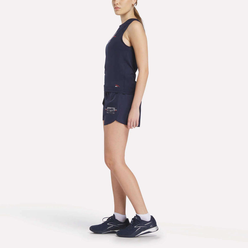 A woman stands sideways in a Women's Reebok x F45 Training Coach Tank Top and matching shorts, both in navy sweat-wicking fabric, paired with white socks and navy sneakers, set against a plain white background.