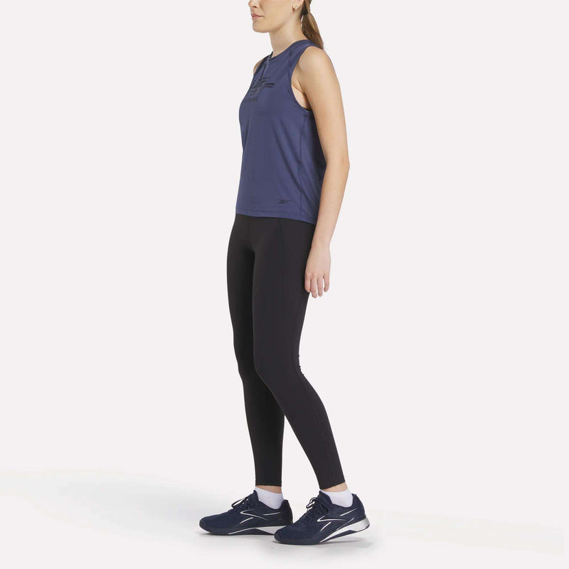 A woman stands slightly sideways in a Women’s Reebok x F45 Training Tank Top, black leggings, and dark blue running shoes against a white background—perfect for F45 workouts.