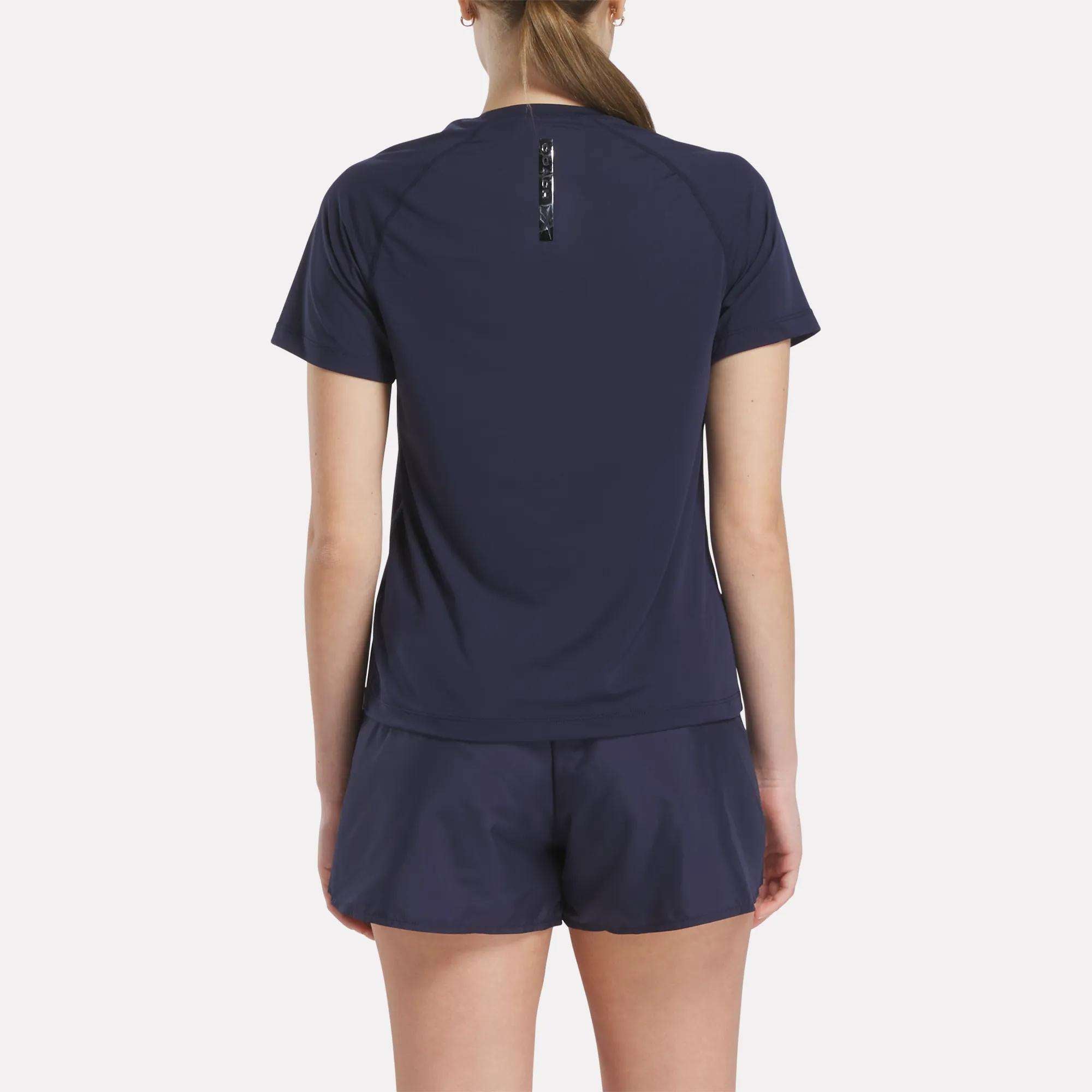 A woman with her hair in a ponytail faces away from the camera, wearing a navy Women's Reebok x F45 Training Coach T-Shirt and matching shorts against a plain white background—ideal for high-intensity workouts.