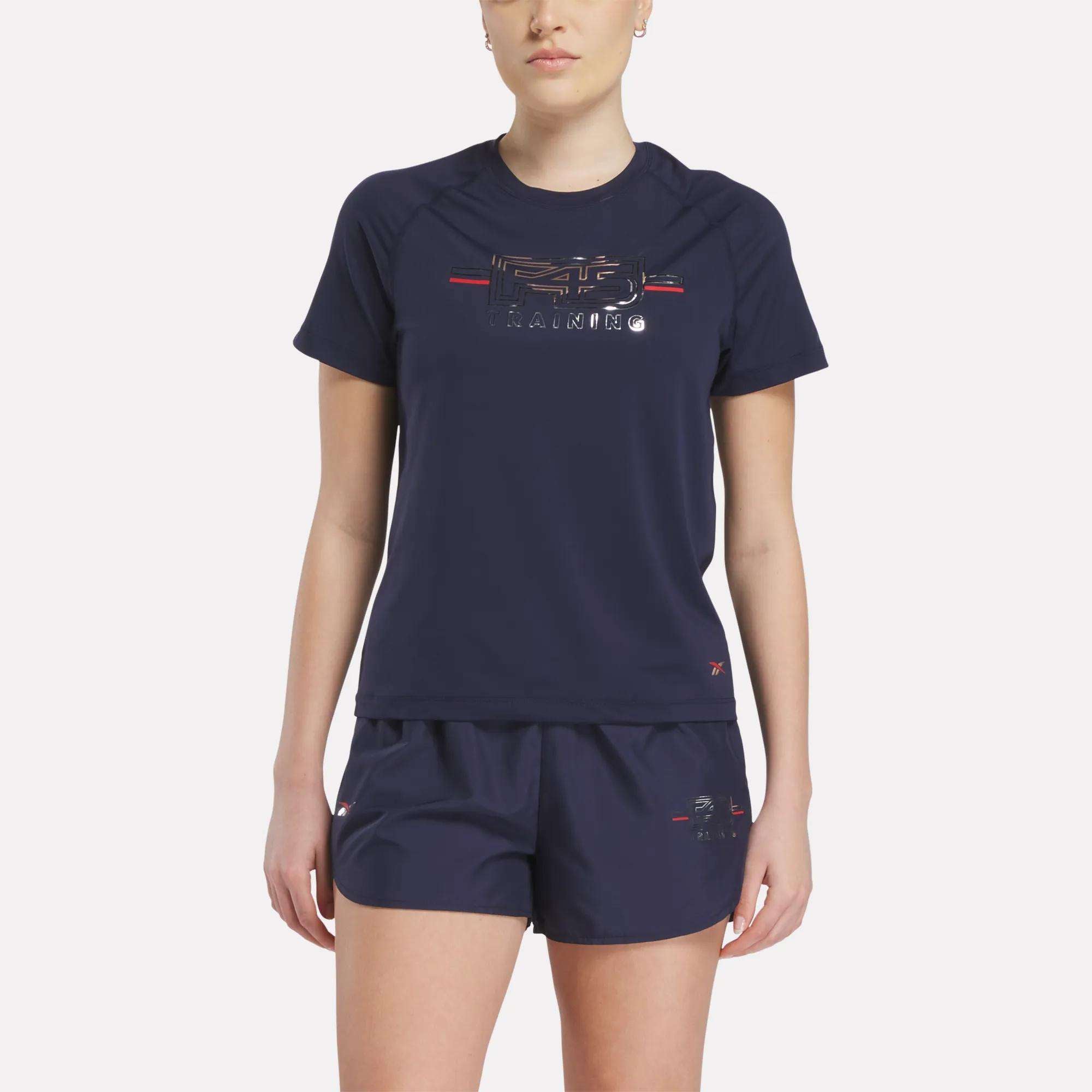 A woman wears a navy Women's Reebok x F45 Training Coach T-Shirt with matching shorts, ideal for high-intensity workouts, as she stands against a plain light background.
