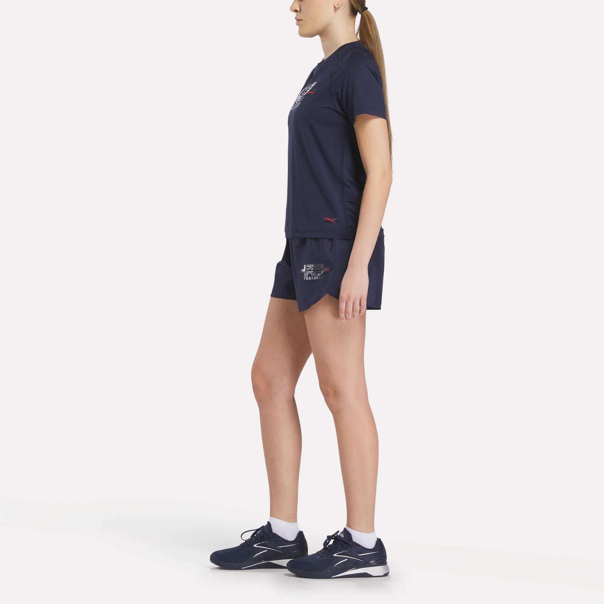 A woman stands in profile against a plain white background, wearing a Reebok Women's Reebok x F45 Training Coach T-Shirt in navy blue, matching shorts, and navy athletic shoes with white soles, ready for high-intensity training.