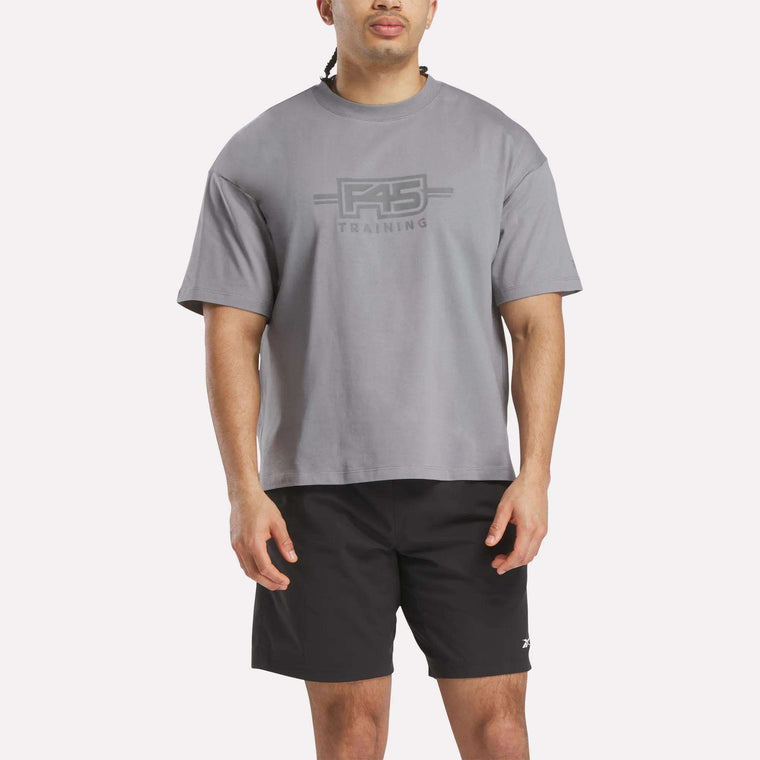 A person wears a loose gray Reebok x F45 Training Tonal Graphic T-Shirt and black shorts, standing facing forward against a plain white background.