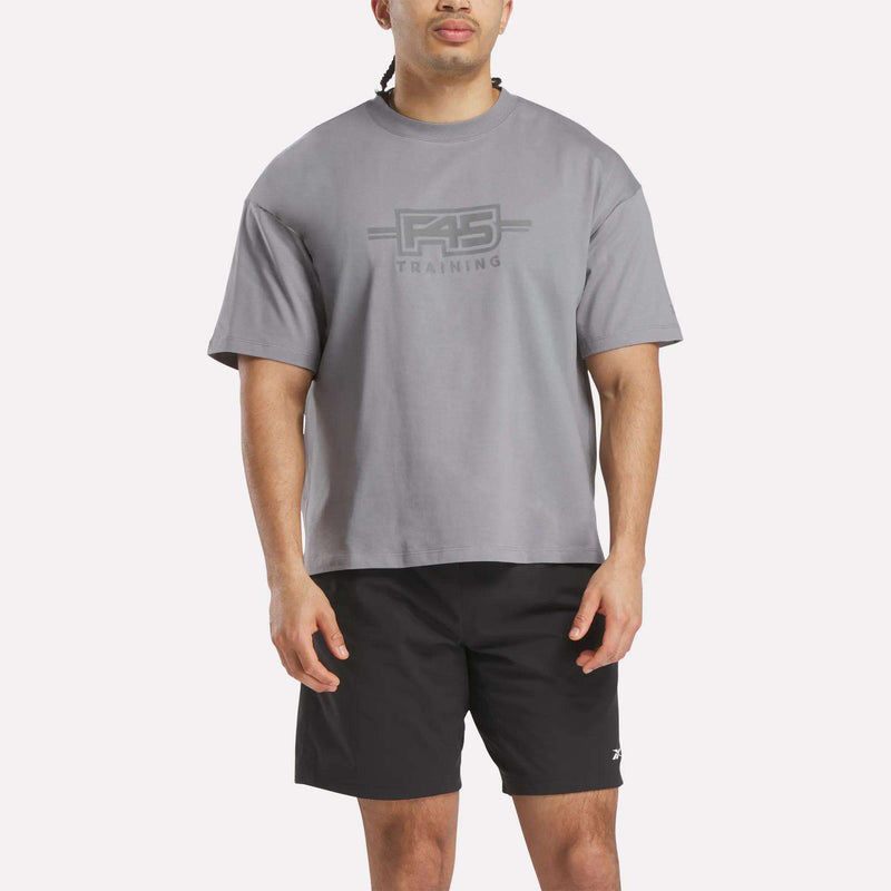 A person wears a loose gray Reebok x F45 Training Tonal Graphic T-Shirt and black shorts, standing facing forward against a plain white background.