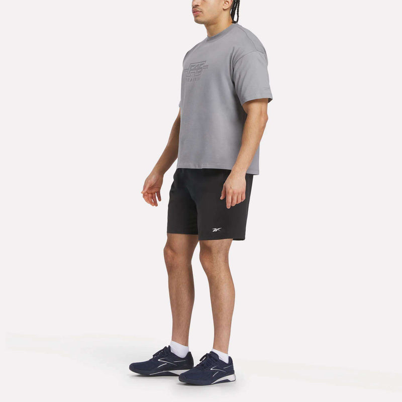 A man stands slightly to the side against a white background, wearing the Reebok x F45 Training Tonal Graphic T-Shirt in gray, black shorts, white socks, and navy blue athletic shoes.