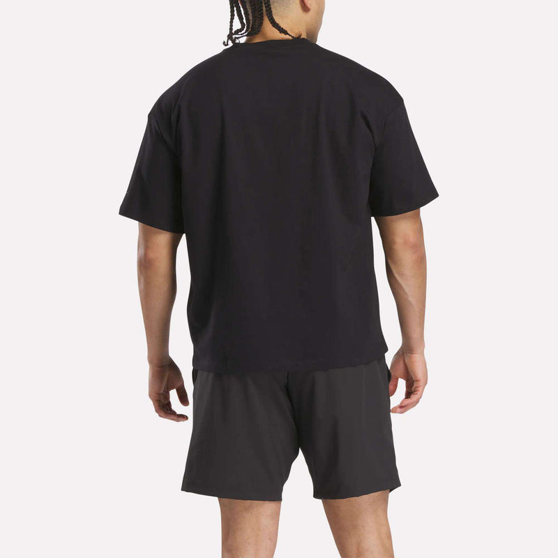 A person with braided hair faces away from the camera, wearing a loose black Reebok x F45 Training Tonal Graphic T-Shirt and black shorts against a plain white background.