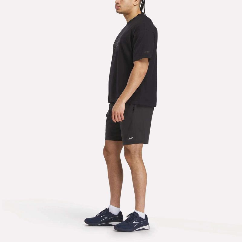 A man stands sideways against a plain white background wearing a Reebok x F45 Training Tonal Graphic T-Shirt, black athletic shorts, white socks, and dark blue sneakers.