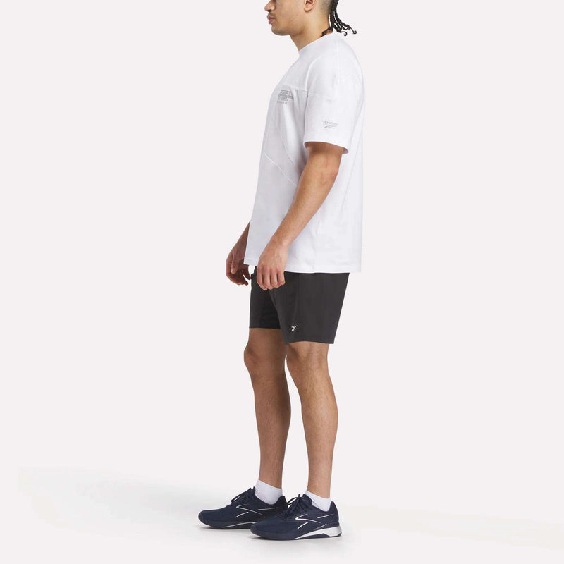 A person stands sideways against a plain, light background, wearing the Reebok x F45 Training Relaxed T-Shirt, black shorts, and navy blue Reebok performance athletic shoes.