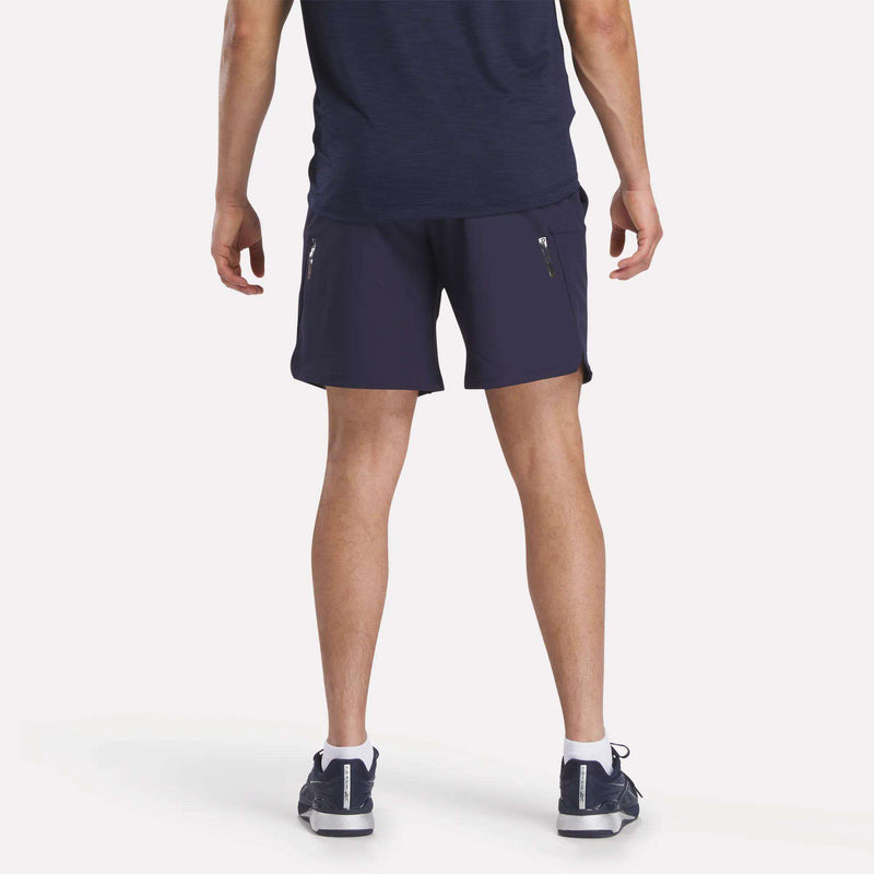 A person stands facing away, wearing a Reebok Men's Long Sleeve Golf Sweatshirt in navy blue with athletic shorts and navy sneakers with white soles, all set against a plain white background.