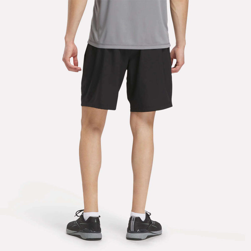 A person dressed in a gray shirt, white socks, black sneakers, and Men's Reebok x F45 Training Woven Shorts is standing with their back to the camera against a plain white background, ready for some high-intensity training.