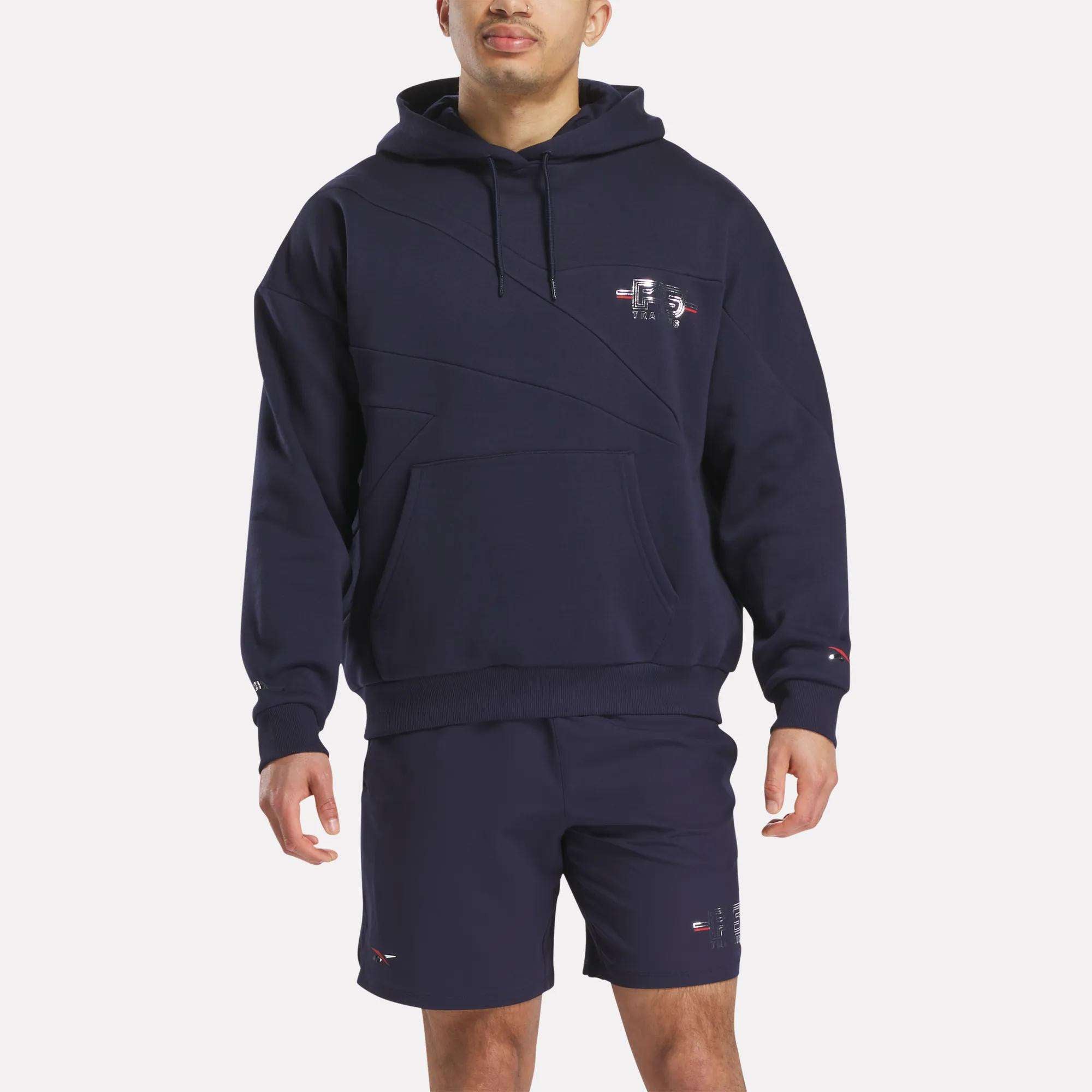 A man faces forward in a navy Reebok x F45 Training Coach Fleece Hoodie with front pocket, subtle seam details, and small logos on chest and sleeve, paired with matching navy shorts. The background is plain white.