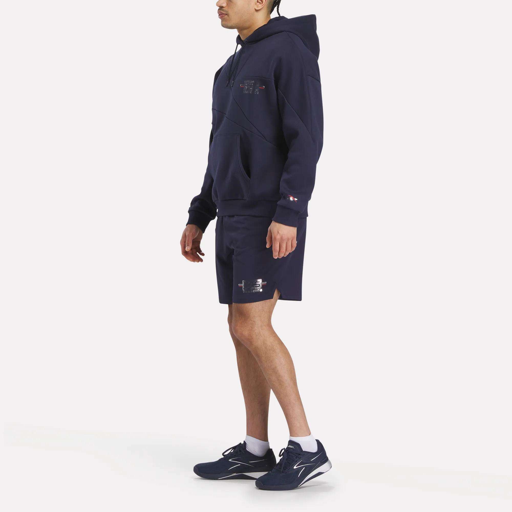 A person stands against a plain white background, facing slightly to the side, wearing the Men's Reebok x F45 Training Coach Fleece Hoodie in navy along with matching shorts and navy sneakers featuring small white logos.