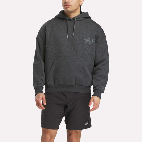 A person wears a Men’s Reebok x F45 Training Relaxed Hoodie in dark gray with a front pocket and black athletic shorts, standing against a plain white background.