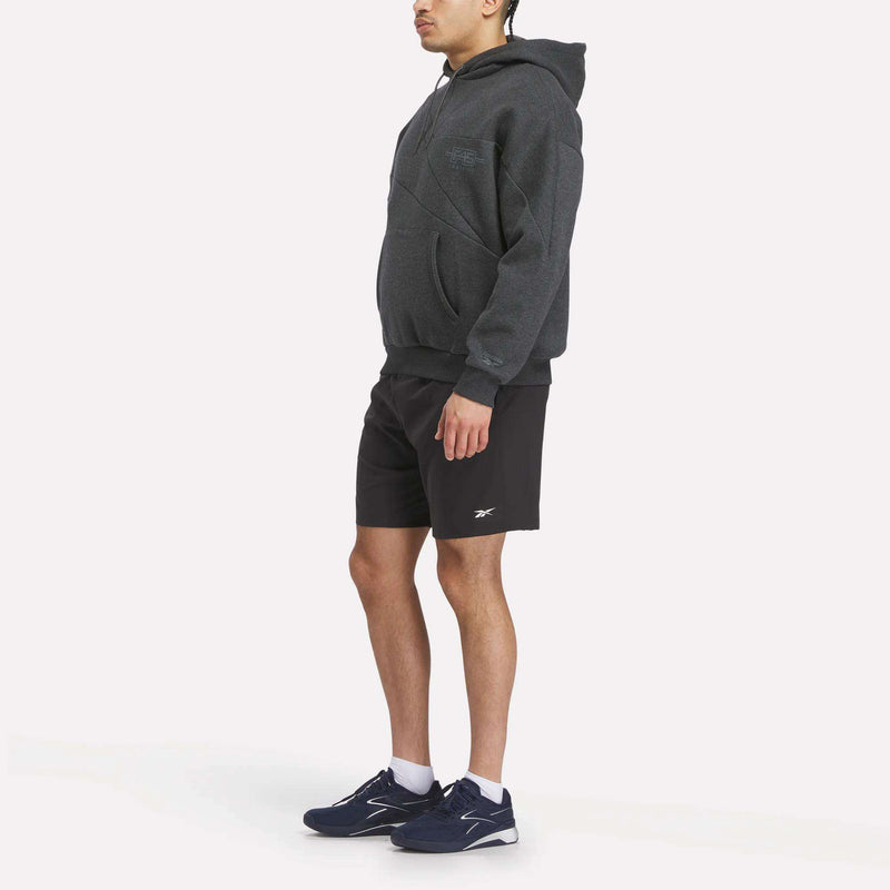 A man stands at an angle against a white background, dressed in the Men's Reebok x F45 Training Relaxed Hoodie in dark gray, black Reebok shorts, and navy blue athletic shoes.