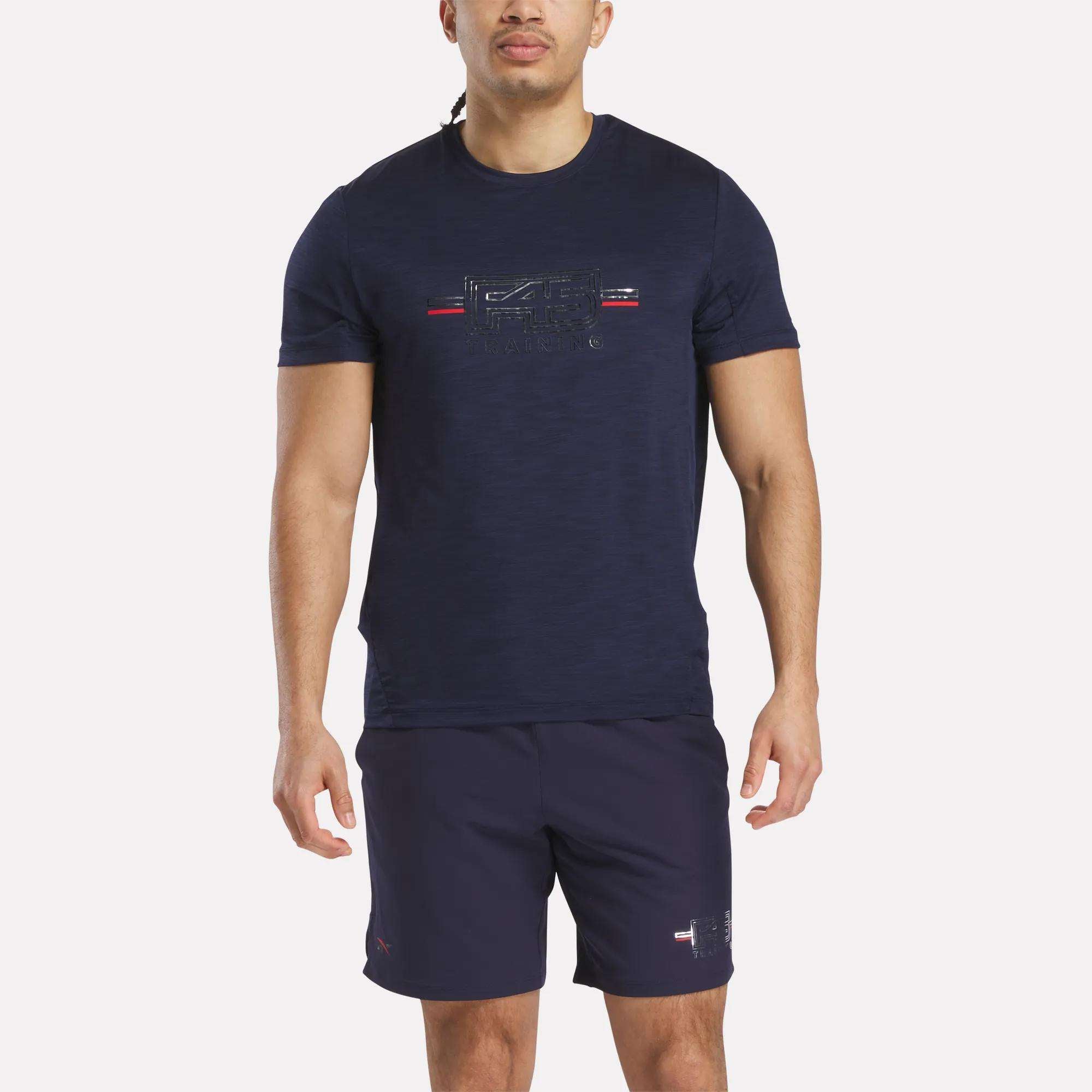A man stands facing forward against a white background, wearing a navy blue Reebok x F45 Training Coach T-Shirt with a small chest graphic, paired with matching navy blue shorts.