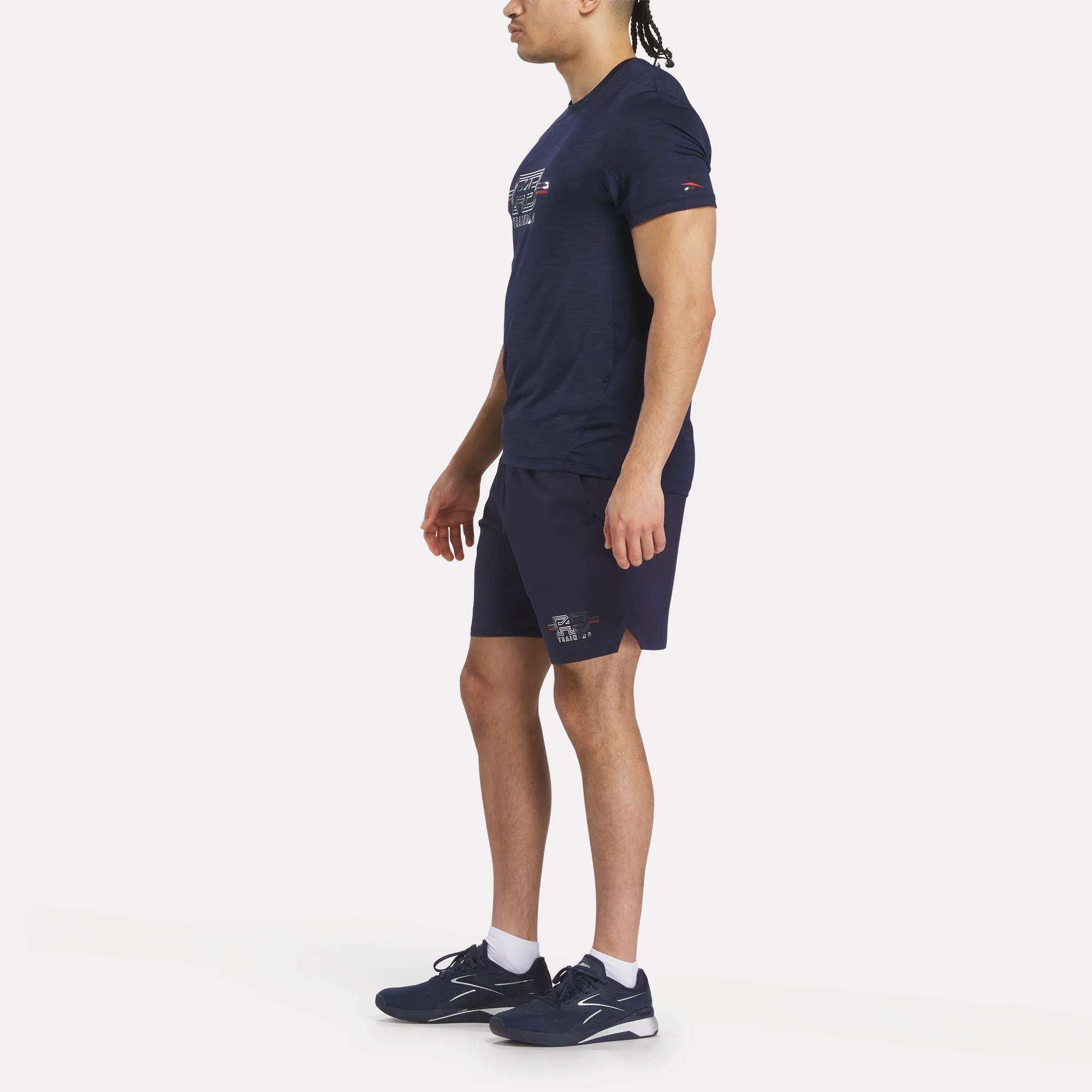 A man stands sideways on a plain white background, ready for an F45 Training session in a navy Men's Reebok x F45 Training Coach T-Shirt, matching shorts, and black sneakers with white soles.